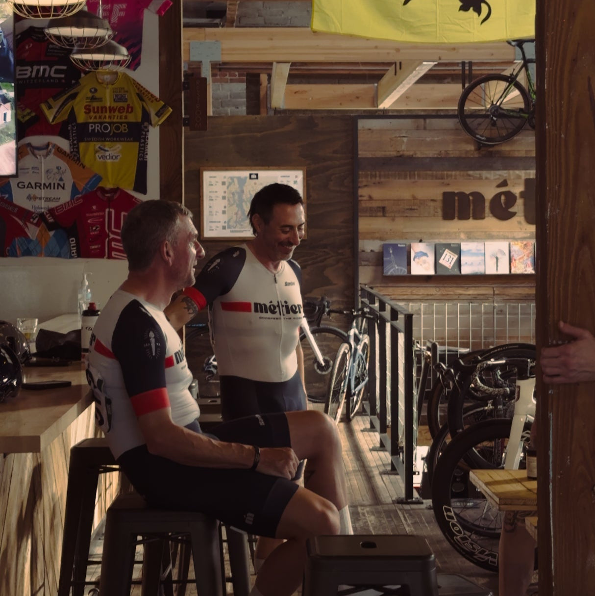 Two Metier Seattle cyclists in Metier team kit sitting at the bar in Metier with cycling-themed decor.